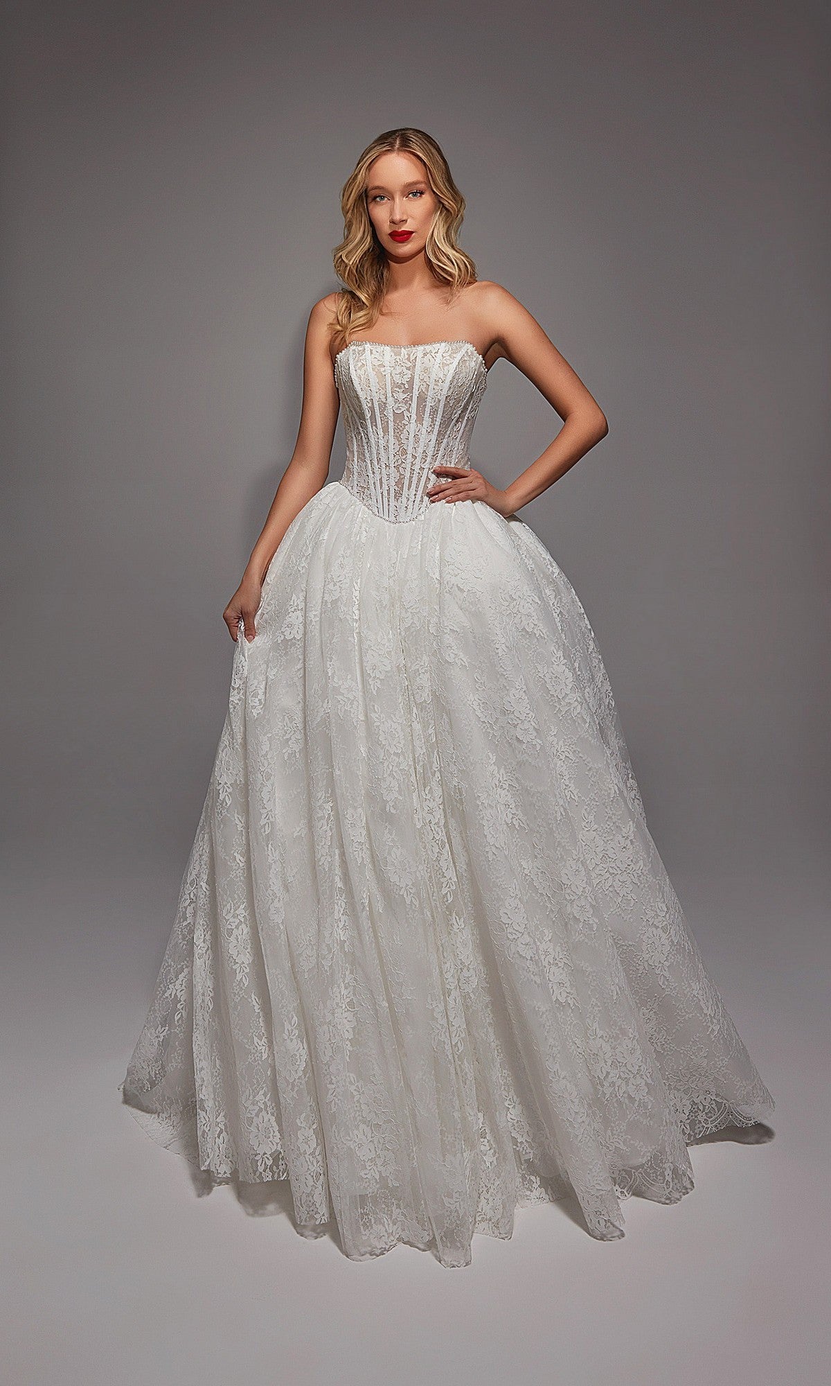 A woman stands in a studio wearing the Long White Dress: Alyce 7149 in Ivory/Sand, featuring a strapless fitted bodice, delicate lace, and full skirt, with one hand on her hip.